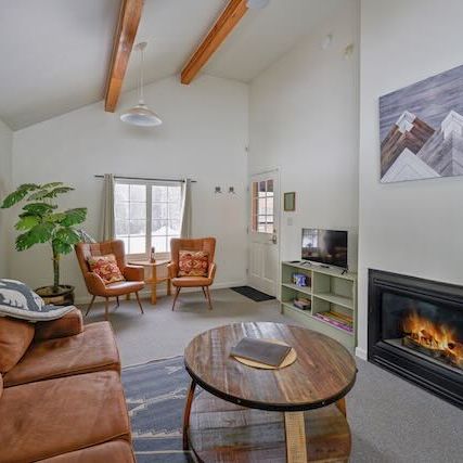 Cozy living room with a brown leather sofa, two orange chairs, wooden coffee table, a fireplace, a flat-screen TV stand, potted plant, and vaulted ceiling with exposed wooden beams.