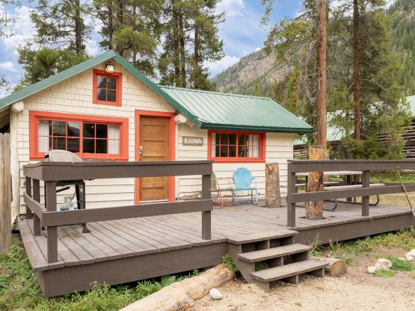 A small rustic cabin with a green roof, red trim, and a wooden porch surrounded by trees, with a picnic table and a barbecue grill on the deck.