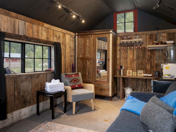 A cozy rustic living space with wood panel walls, a blue sofa, a small chair, a window, and a compact kitchenette area.