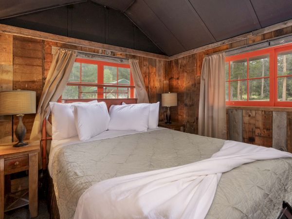 Cozy rustic bedroom with a large bed, cream bedding, wooden paneled walls, red-framed windows, two nightstands, and soft lighting.