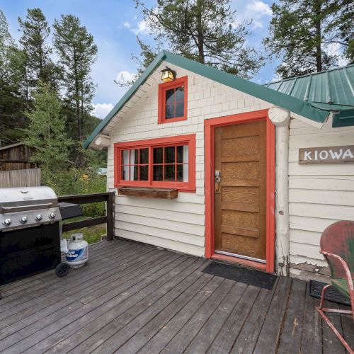 A small, colorful garage with a tan front and red trim sits beside a carport; a wooden fence and trees surround the tidy yard.