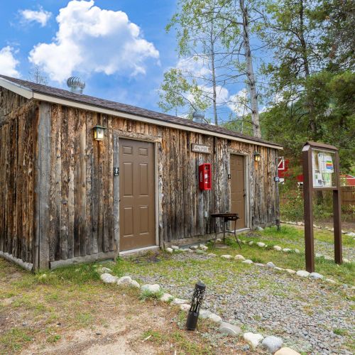 A rustic, weathered wooden cabin with a brown door, fire extinguisher on the wall, gravel path, pine trees, and a blue sky with clouds.