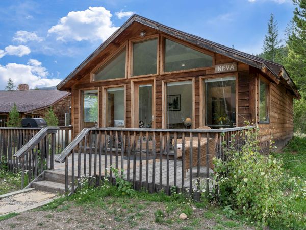 A cozy wooden cabin with large glass windows, a rustic porch, and lush greenery surrounding it, under a partly cloudy blue sky.