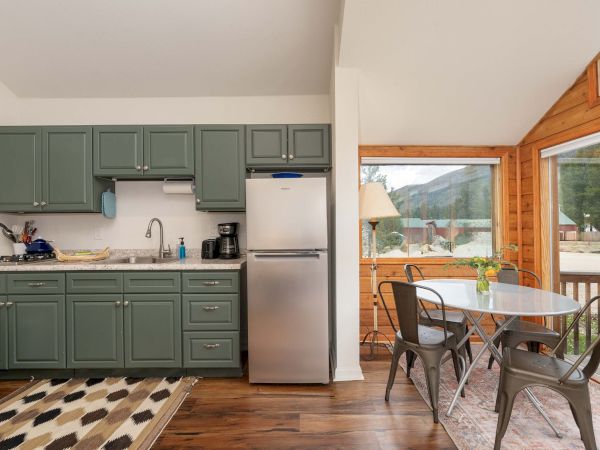 A compact kitchen with sage green cabinets, stainless fridge, and a small dining nook by large windows overlooking a wooden deck.