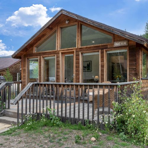 A small wooden cabin with a veranda, set on a hillside, surrounded by a fence and trees, under a clear blue sky.