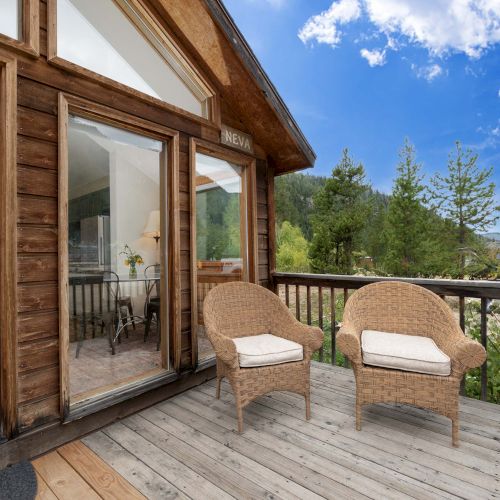 A cozy outdoor patio with wooden beams, a tiled floor, a wooden door, and potted plants under a bright blue sky with a few clouds.