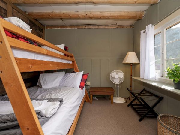 A cozy bunk bedroom with a wooden bunk bed, pillows, a small side table, a fan, a window, a lamp, a plant, and a wicker basket.