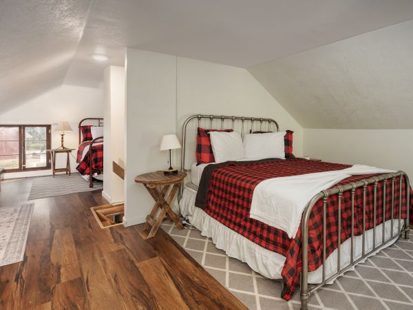 Cozy attic bedroom with a wrought-iron bed, red plaid bedding, two nightstands, and wood floors; a seating area and balcony visible in the background.