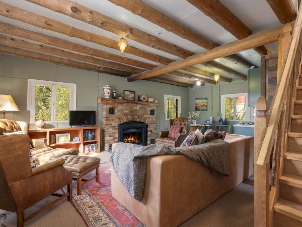 Cozy living room with a stone fireplace, exposed wooden beams, and comfy seating arranged around a rug and TV, nearby stairs and windows.