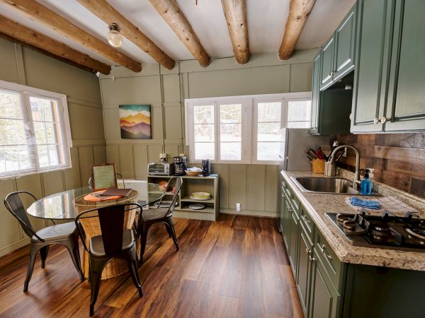 Cozy kitchen with green cabinets, a round dining table, four chairs, wooden beams, and a bright windowed wall, wood floors, and a small art piece on the wall.