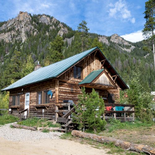 A cozy wooden cabin nestled in tall pines, with a sloped roof, stone chimney, and a sunny mountain backdrop.