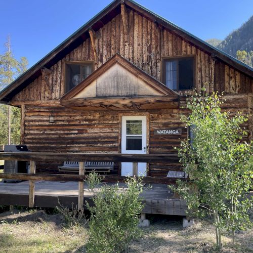 A rustic wooden cabin with a peaked roof, two stories, surrounded by pine trees and a small front porch.