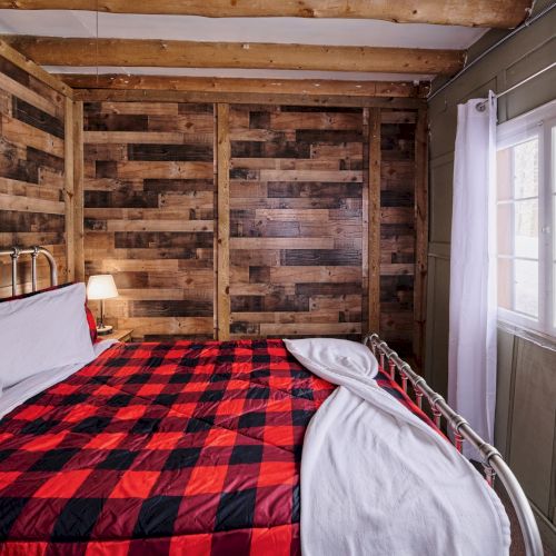 Cozy rustic bedroom with a metal-framed bed, red and black plaid blanket, stacked wood plank walls, bedside lamp, and white curtains by a window.