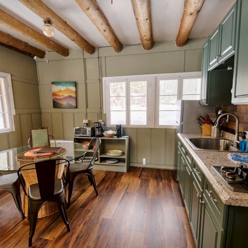 Cozy kitchen-dining area with green cabinets, wooden beams, a small round table and chairs, windows, and a sleek countertop stove.