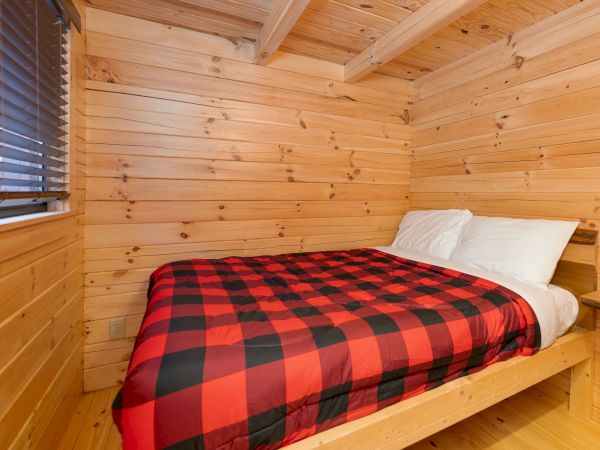 A small wooden-paneled bedroom with a double bed, white pillows, and a red-and-black buffalo plaid blanket, cozy and rustic.