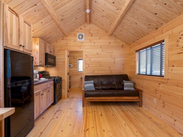 A cozy wooden cabin interior with a kitchen on the left, a black sofa center, wood-paneled walls and ceiling, and a window on the right, all in warm tones.