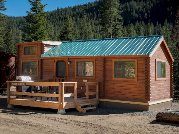 A small log cabin with a teal metal roof, wooden deck, and surrounding forest; sunlight, trees, and a clear blue sky in a mountain setting. ending punctuation.