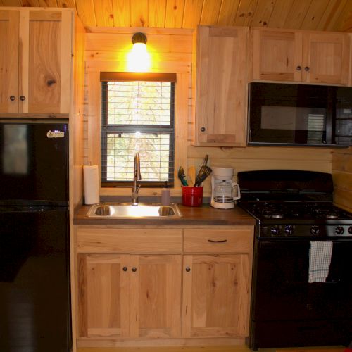 A compact wooden kitchen with light timber cabinets, black refrigerator, microwave, stove, sink under a window, and a small counter area.