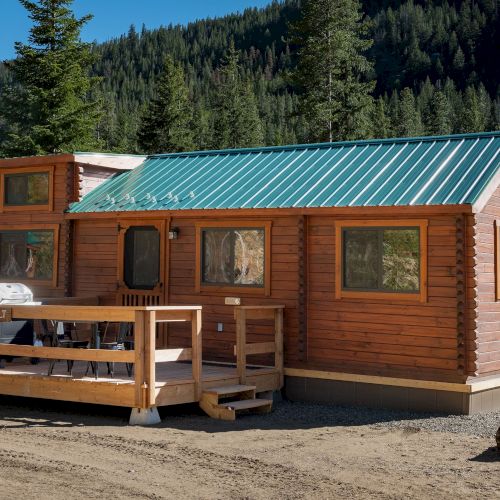 A small log cabin with a teal metal roof sits in a forest clearing, featuring a wooden deck and large windows.