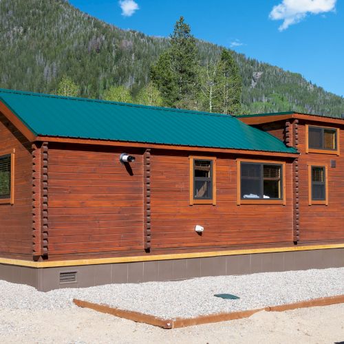 A small wooden cabin with a green metal roof sits on a concrete foundation, surrounded by trees and mountains under a blue sky.