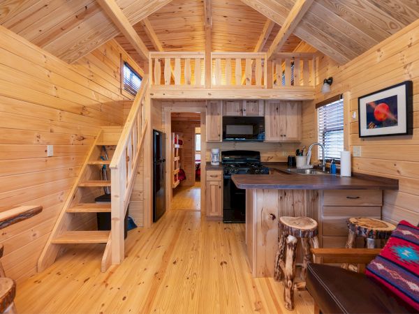 Cozy log cabin interior with a loft, wooden stairs, compact kitchen, and rustic furniture; warm wood tones, a small dining nook, and framed art on the wall.