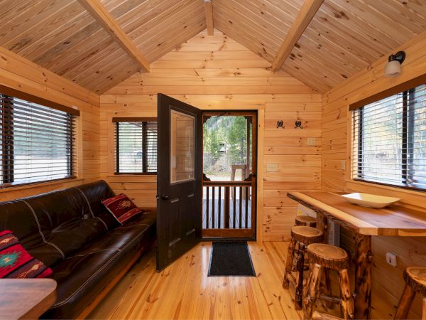 Cozy wooden cabin interior with a black couch, open front door to a deck, large windows with blinds, and a small counter with stools along the right wall.