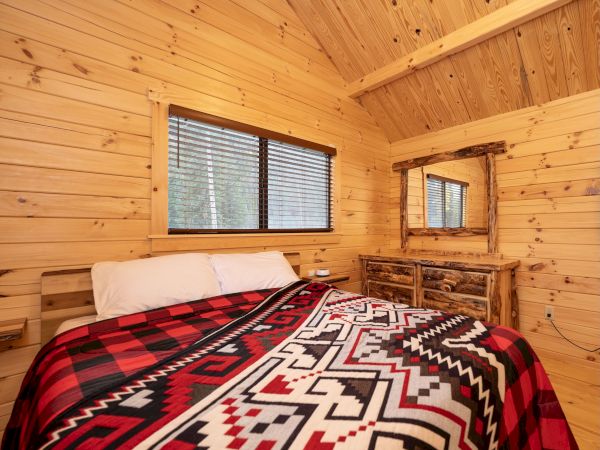 Cozy wooden cabin bedroom with a patterned red-black blanket, mirror, window, and wood furniture. Sounds like a rustic retreat.