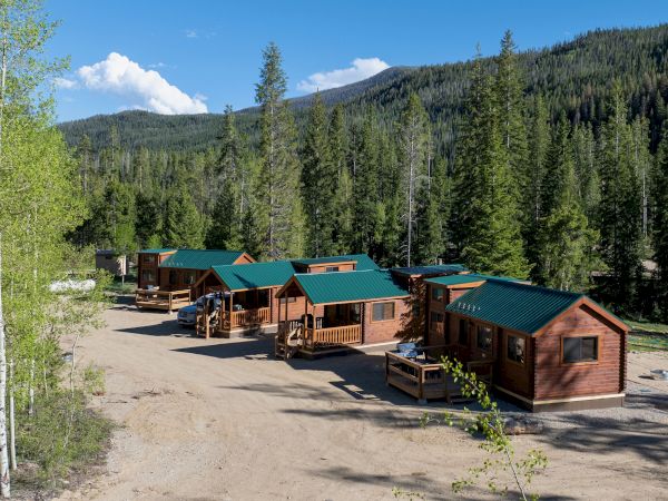 Mini wooden cabins with green roofs line a clearing, set among tall pine trees and forested hills under a blue sky, a peaceful campsite.