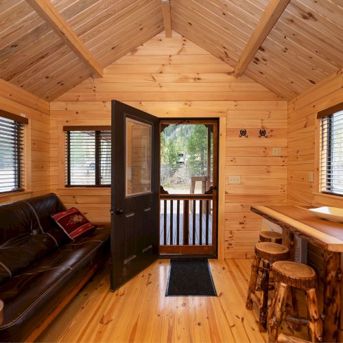 A cozy wooden cabin interior with a black sofa, open door to a deck, multiple windows with blinds, and a wooden bar with stools on the right.