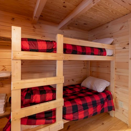 A wooden cabin bedroom with a bunk bed set: two top and bottom bunks in red-and-black plaid, stacked against pine walls and floor, plus shelves and another bed in the background.