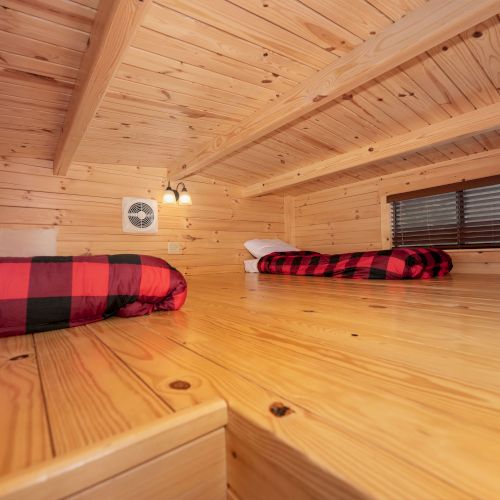 A cozy wooden attic bedroom with two red and black plaid blankets on low beds, natural pine walls, a small ceiling light, and a window with blinds.