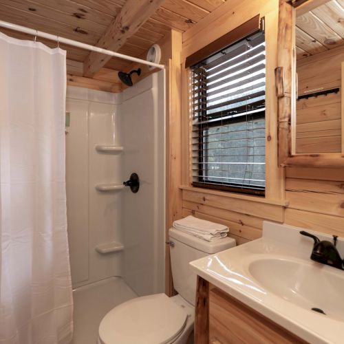 A narrow, modern kitchen with light wood cabinets, a small island, and stainless appliances along a bright corridor ending in a doorway.