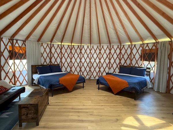 A cozy yurt interior with two single beds, each with blue bedding and orange throws, wooden floor, lattice walls, and natural light.