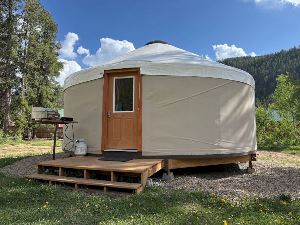 A beige yurt with a wooden door sits on a small deck, set on rocky ground amid trees and mountains under a blue sky.