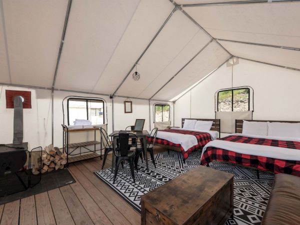 A cozy, rustic tented living space with a wood stove, stacked firewood, a dining table, chairs, and two beds with plaid red blankets, on a wooden deck.