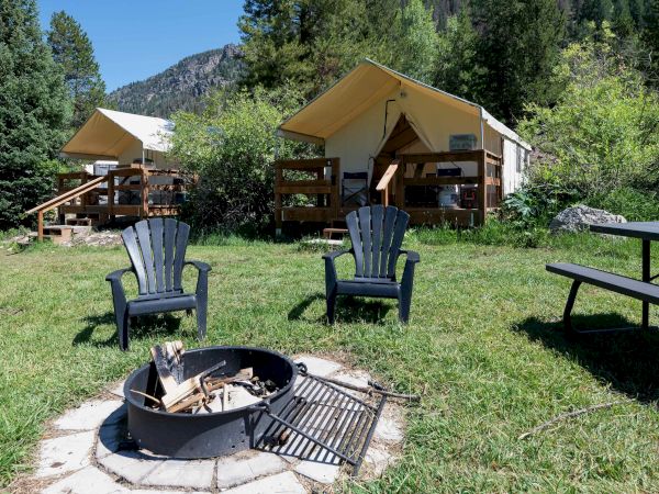 Campground with two canvas cabins, wooden porches, a fire pit, two lawn chairs, a picnic table, tall trees, and a bright sunny sky.