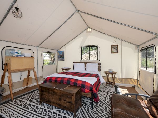 A spacious tent bedroom with a queen bed, plaid blanket, wooden crate coffee table, vintage dresser, rug, sofa, side tables, and windows.