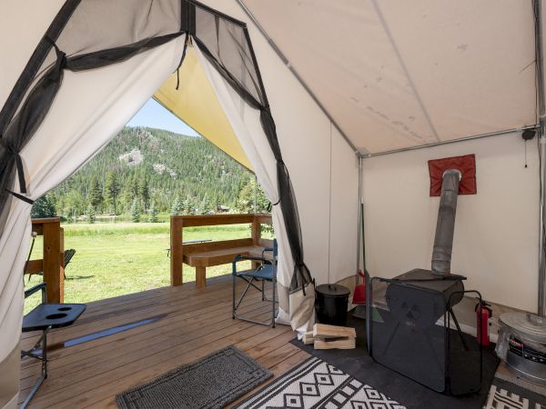 Cozy tent interior with a wood stove, hanging coordinates, rug, and a view of green grass and trees through the open flap, inviting outdoor vibes.