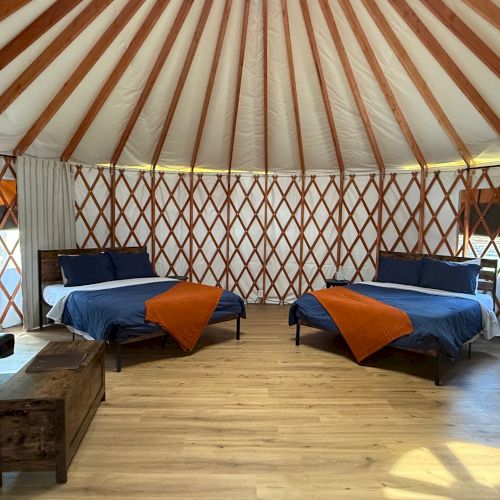 A spacious yurt-style room with wooden beams, two blue-bed cabins, orange throws, a wooden floor, and cozy seating&mdash;bright, airy, and inviting.