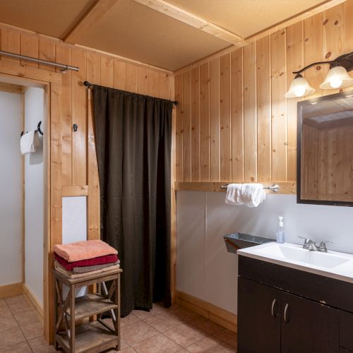 A rustic bathroom with wooden panel walls, a dark vanity with a white sink, framed mirror, two light fixtures, and a small seating area with towels; curtain partitions + entryway.