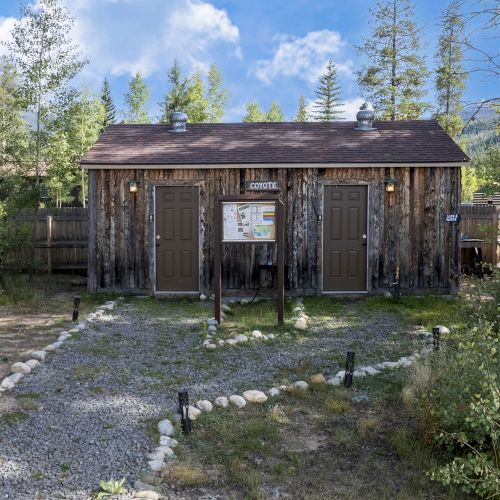 A rustic wooden restroom building in a forest clearing, with gravel path, trees, and a small sign in front ending here.
