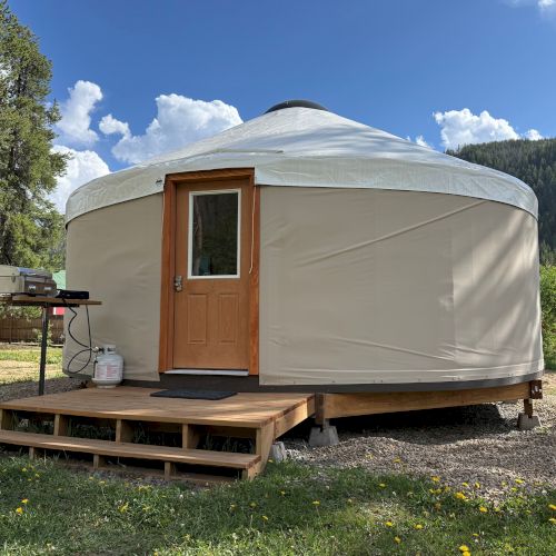 A round, canvas yurt on a wooden platform with a small front porch, door, grill nearby, set on grassy, wooded land under blue sky.