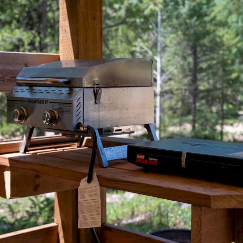 A small gas grill and a portable black case sit on a wooden deck railing, overlooking a forested outdoor area.