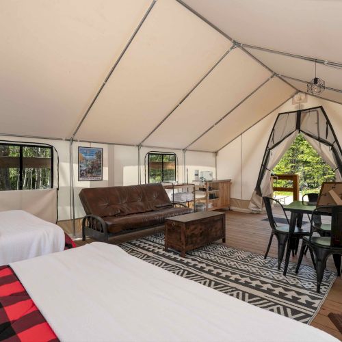 A large, bright glamping tent with two beds, a sofa, coffee table, rug, and a dining area with black chairs and a geometric tent nook, outside view visible.