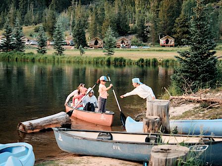 A lakeside scene with people in a small orange boat, one person paddling, others aboard or nearby, wooden docks, pine trees, and a forested shore with cabins in the background.