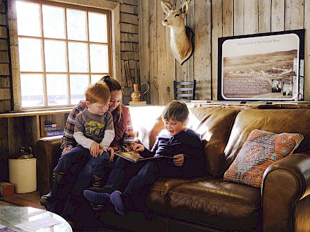 Three kids read together on a brown sofa in a cozy cabin, sunlight streaming from a window, a mounted deer head, and vintage decor nearby.
