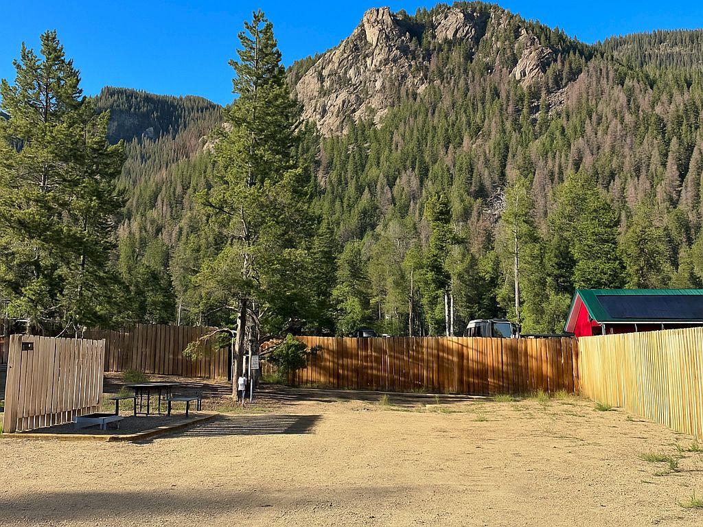 A sunny mountain yard with a wooden fence, pine trees, and rugged cliffs in the background over a dry, dirt ground.