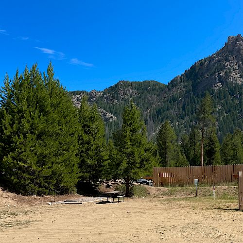 A sunny mountain landscape with tall pines, rocky hills, and a sandy open area; a wooden fence lines the right, under a bright blue sky.