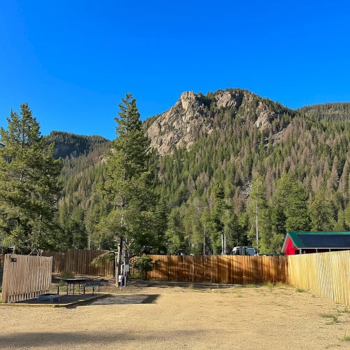 A desert-like landscape with rocky cliffs, clear blue sky, and a few trees near a rustic wooden fence and bench, creating a sunny, arid scene.