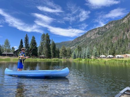 A person stands on a blue canoe in calm lake water, surrounded by pine trees, mountains, and small cabins under a bright, partly cloudy sky.